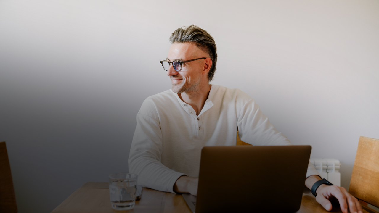 Ein älterer Mann mit Brille sitzt vor einem Laptop und blickt lächelnd zur Seite.
